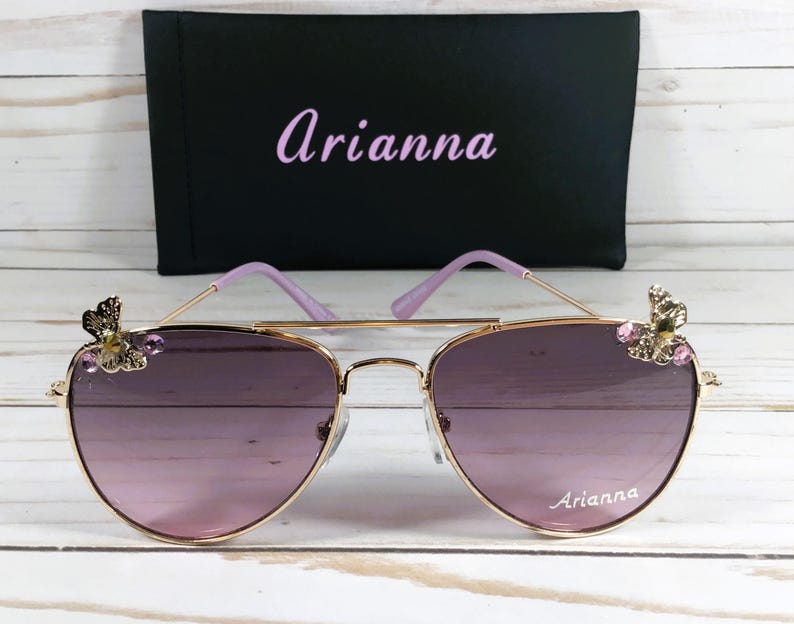 Op de afbeelding: Gouden aviator zonnebril met paarse glazen en vlinderversieringen op de pootjes. De zonnebril heeft de naam "Arianna" op het rechter glas.