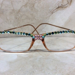 May include: Gold-toned eyeglasses with clear lenses, adorned with pink, green, and blue rhinestones along the top and corners of the frames. The glasses have a classic rectangular shape with a delicate wire frame.