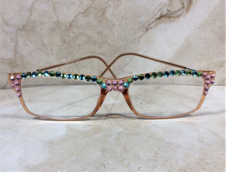 May include: Gold-toned eyeglasses with clear lenses, adorned with pink, green, and blue rhinestones along the top and corners of the frames. The glasses have a classic rectangular shape with a delicate wire frame.