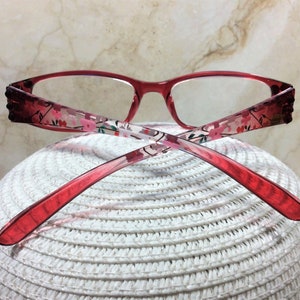 Puede incluir: Gafas con montura roja y estampado floral en las varillas. Las gafas tienen forma rectangular y descansan sobre una superficie blanca tejida. Las varillas presentan un degradado de rojo oscuro a claro.