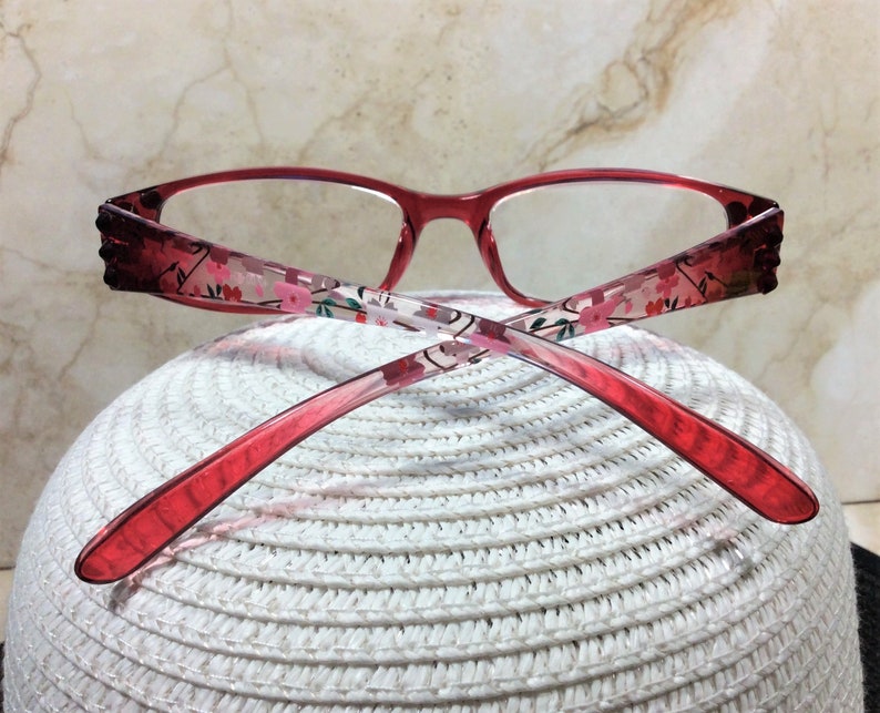 Puede incluir: Gafas con montura roja y estampado floral en las varillas. Las gafas tienen forma rectangular y descansan sobre una superficie blanca tejida. Las varillas presentan un degradado de rojo oscuro a claro.