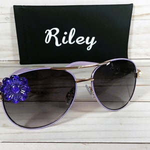 Pu&ograve; includere: Occhiali da sole aviator viola con montatura dorata e un fiorellino blu come decorazione sul lato sinistro.