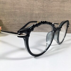 Puede incluir: Gafas negras con lentes transparentes y una montura adornada con peque&ntilde;os cristales negros facetados en el borde superior. Las patillas est&aacute;n fabricadas con una combinaci&oacute;n de material negro y componentes de metal plateado pulido.