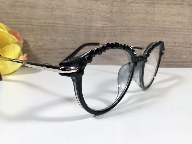 Puede incluir: Gafas negras con lentes transparentes y una montura adornada con peque&ntilde;os cristales negros facetados en el borde superior. Las patillas est&aacute;n fabricadas con una combinaci&oacute;n de material negro y componentes de metal plateado pulido.