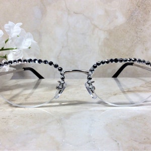 Pu&ograve; includere: Occhiali con montatura argentata e lenti trasparenti. I bordi superiori sono impreziositi da una fila di piccole strass grigio scuro. Gli occhiali hanno un design a mezza montatura e aste nere. Lo stile generale &egrave; elegante e decorativo.