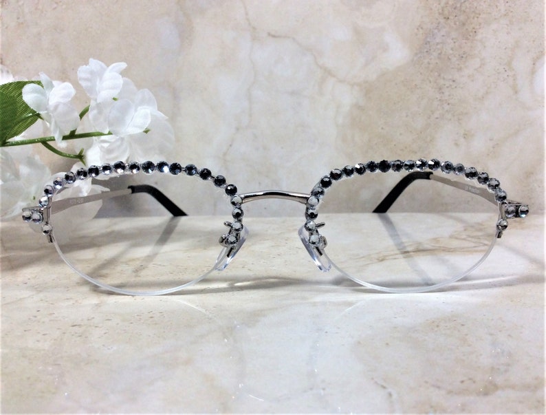 Pu&ograve; includere: Occhiali con montatura argentata e lenti trasparenti. I bordi superiori sono impreziositi da una fila di piccole strass grigio scuro. Gli occhiali hanno un design a mezza montatura e aste nere. Lo stile generale &egrave; elegante e decorativo.