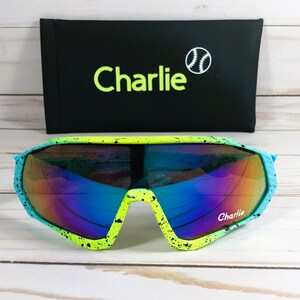 Puede incluir: Gafas de sol verdes y azules con un dise&ntilde;o de pintura salpicada en blanco y negro. Las gafas de sol tienen un estilo envolvente y dicen "Charlie" en la lente derecha. Las gafas de sol est&aacute;n en una caja negra con la palabra "Charlie" y un gr&aacute;fico de b&eacute;isbol.