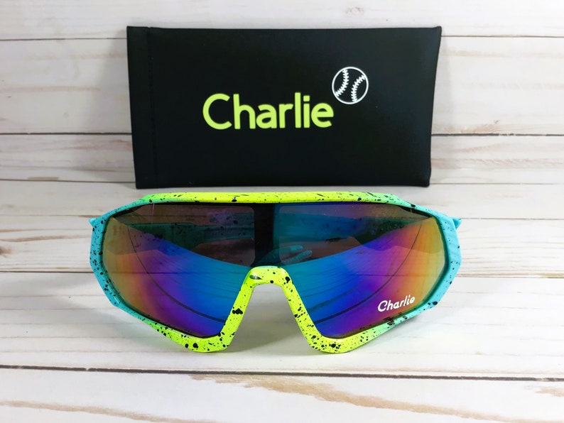Puede incluir: Gafas de sol verdes y azules con un dise&ntilde;o de pintura salpicada en blanco y negro. Las gafas de sol tienen un estilo envolvente y dicen "Charlie" en la lente derecha. Las gafas de sol est&aacute;n en una caja negra con la palabra "Charlie" y un gr&aacute;fico de b&eacute;isbol.