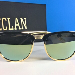 May include: Black sunglasses with gold accents and green mirrored lenses. The sunglasses are on a white surface.