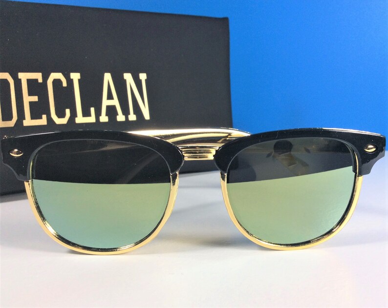 May include: Black sunglasses with gold accents and green mirrored lenses. The sunglasses are on a white surface.