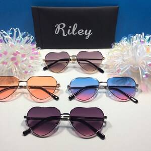 May include: Five pairs of heart-shaped sunglasses in different colors, including pink, blue, and brown. The sunglasses are arranged in a row on a white surface. The word "Riley" is printed on a black case in the background.