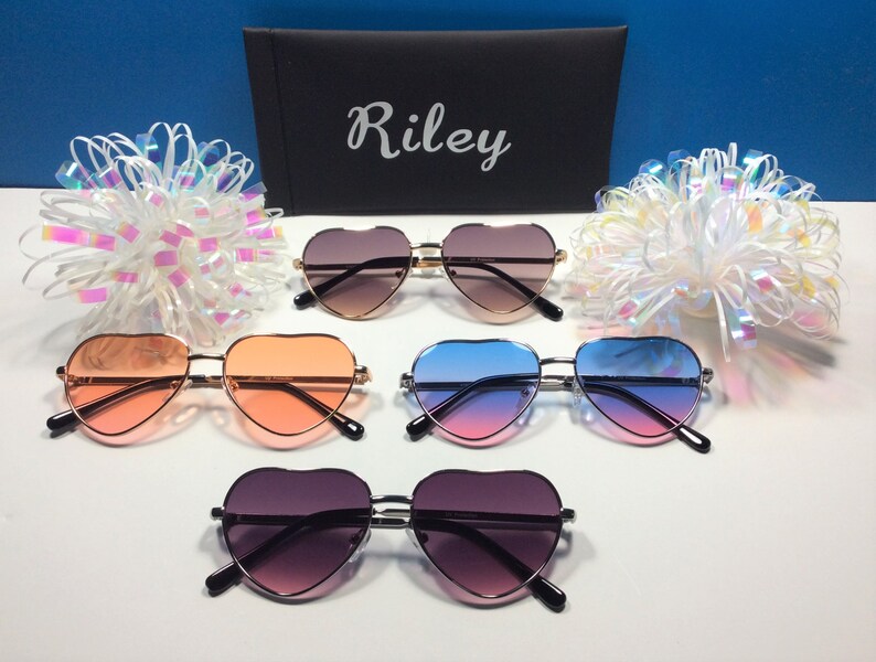 May include: Five pairs of heart-shaped sunglasses in different colors, including pink, blue, and brown. The sunglasses are arranged in a row on a white surface. The word "Riley" is printed on a black case in the background.