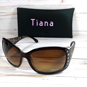 K&ouml;nnte beinhalten: Braune und schwarze Sonnenbrille mit Strasssteinen an der Seite. Die Sonnenbrille hat braune Gl&auml;ser und einen schwarzen Rahmen. Die Sonnenbrille befindet sich in einem schwarzen Etui mit dem aufgedruckten Wort "Tiana".