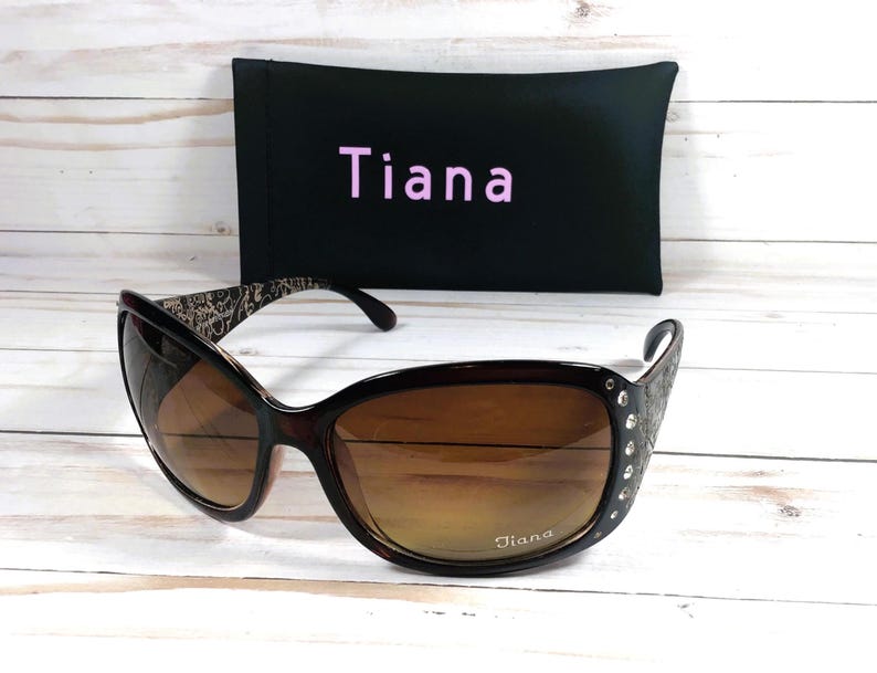 K&ouml;nnte beinhalten: Braune und schwarze Sonnenbrille mit Strasssteinen an der Seite. Die Sonnenbrille hat braune Gl&auml;ser und einen schwarzen Rahmen. Die Sonnenbrille befindet sich in einem schwarzen Etui mit dem aufgedruckten Wort "Tiana".