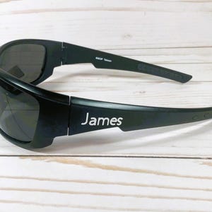 Puede incluir: Gafas de sol envolventes negras con lentes oscuras y montura negra mate. La patilla muestra el nombre James impreso en blanco, mientras que la patilla interior incluye el texto 8263P Taiwan en letras peque&ntilde;as de color blanco.