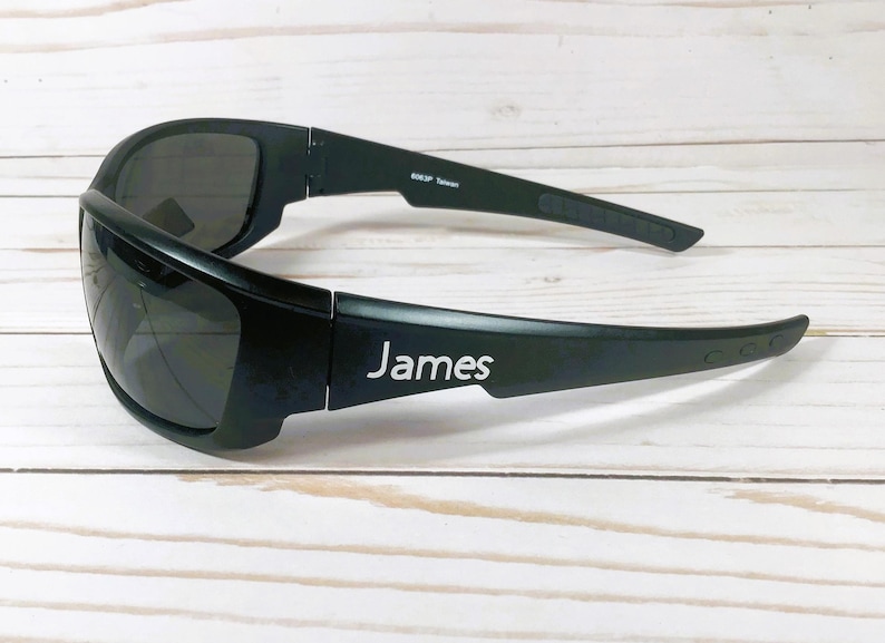 Puede incluir: Gafas de sol envolventes negras con lentes oscuras y montura negra mate. La patilla muestra el nombre James impreso en blanco, mientras que la patilla interior incluye el texto 8263P Taiwan en letras peque&ntilde;as de color blanco.