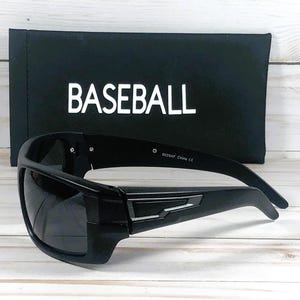 Pu&ograve; includere: Occhiali da sole neri con montatura rettangolare e lenti scure. Gli occhiali sono davanti a un astuccio nero con la scritta "BASEBALL" stampata in bianco. Lo sfondo &egrave; un legno chiaro.
