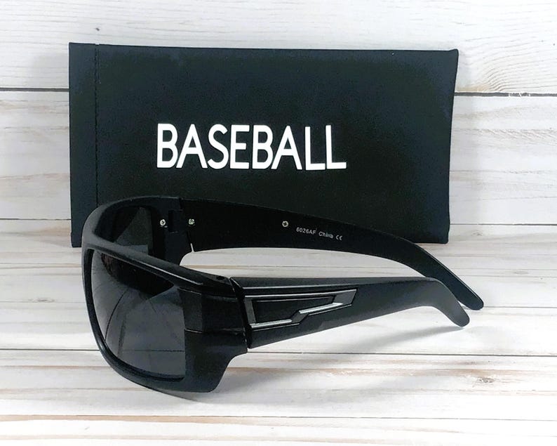 Pu&ograve; includere: Occhiali da sole neri con montatura rettangolare e lenti scure. Gli occhiali sono davanti a un astuccio nero con la scritta "BASEBALL" stampata in bianco. Lo sfondo &egrave; un legno chiaro.