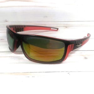May include: Black and red sunglasses with a rectangular frame. The lenses have a gradient tint, transitioning from green to yellow. The temples are black with a decorative emblem.