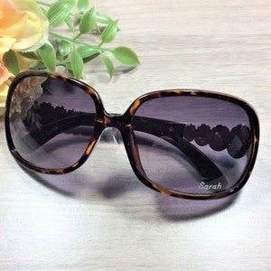 Puede incluir: Gafas de sol de carey con lentes oscuros y un acento de cuentas en el lateral. Las gafas de sol tienen la palabra "Sarah" impresa en la lente derecha.