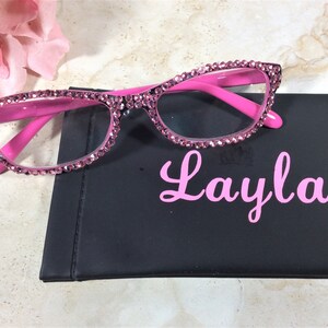 Pu&ograve; includere: Occhiali rosa con strass e montatura rosa abbinata appoggiati su un astuccio nero. Sull'astuccio &egrave; stampato in rosa il nome "Layla". Sullo sfondo c'&egrave; un fiore rosa.