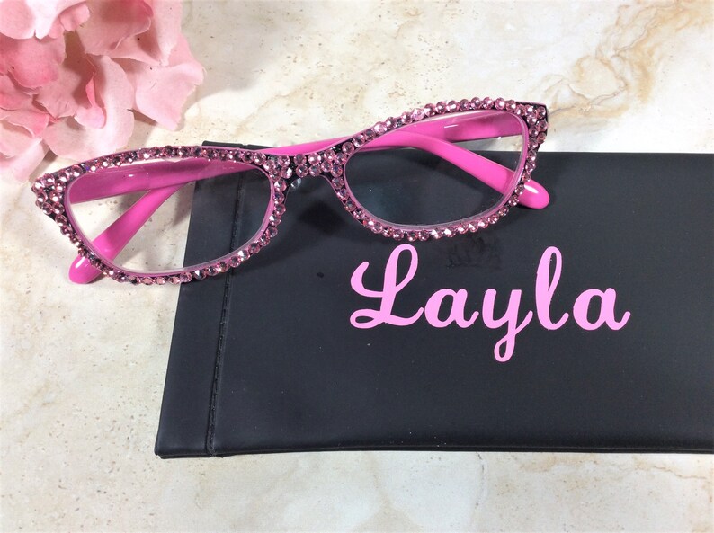 Pu&ograve; includere: Occhiali rosa con strass e montatura rosa abbinata appoggiati su un astuccio nero. Sull'astuccio &egrave; stampato in rosa il nome "Layla". Sullo sfondo c'&egrave; un fiore rosa.
