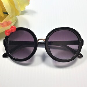 May include: Black round sunglasses with a red bow on the left side. The sunglasses have a gold accent on the bridge.