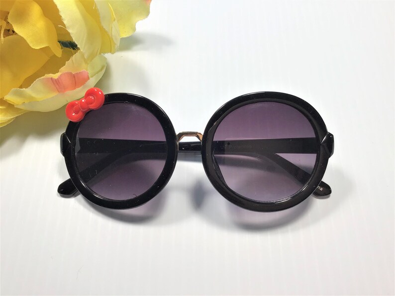 May include: Black round sunglasses with a red bow on the left side. The sunglasses have a gold accent on the bridge.