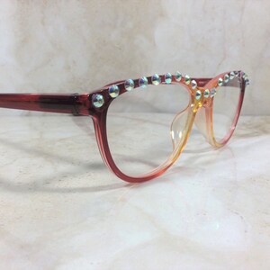 Puede incluir: Gafas con una montura degradada que pasa de rojo oscuro a naranja claro. El borde superior de la montura est&aacute; adornado con una fila de peque&ntilde;os cristales circulares iridiscentes que reflejan la luz con un efecto brillante multicolor.