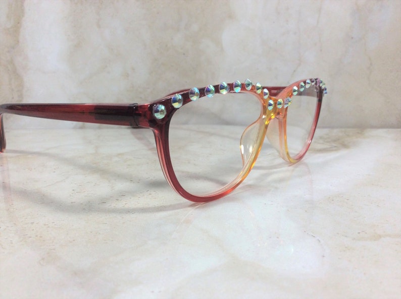 Puede incluir: Gafas con una montura degradada que pasa de rojo oscuro a naranja claro. El borde superior de la montura est&aacute; adornado con una fila de peque&ntilde;os cristales circulares iridiscentes que reflejan la luz con un efecto brillante multicolor.