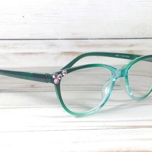 May include: Green gradient eyeglasses featuring a translucent frame that transitions from a dark forest green at the temples to a lighter mint green at the bottom. The outer corners are embellished with small, round, light pink decorative crystals.