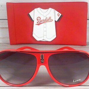 Puede incluir: Gafas de sol rojas de aviador con lentes oscuras y el nombre "Liam" en la lente derecha. Una funda roja con un dise&ntilde;o de camiseta de b&eacute;isbol est&aacute; detr&aacute;s de las gafas.