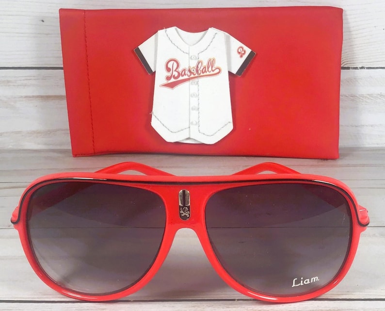 Puede incluir: Gafas de sol rojas de aviador con lentes oscuras y el nombre "Liam" en la lente derecha. Una funda roja con un dise&ntilde;o de camiseta de b&eacute;isbol est&aacute; detr&aacute;s de las gafas.