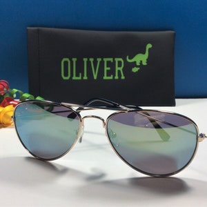 May include: Gold aviator sunglasses with blue mirrored lenses. The sunglasses are in a black case with the word "OLIVER" and a green dinosaur graphic.