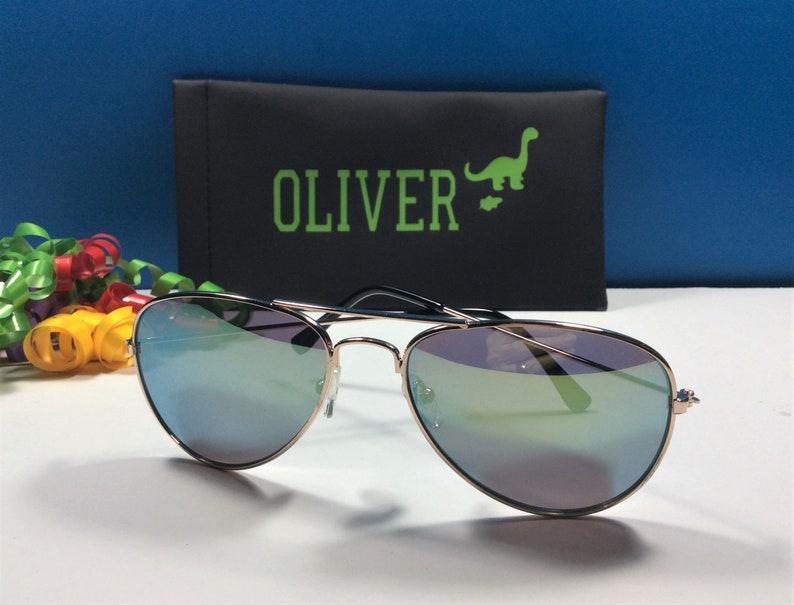 May include: Gold aviator sunglasses with blue mirrored lenses. The sunglasses are in a black case with the word "OLIVER" and a green dinosaur graphic.