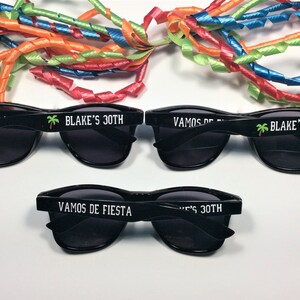 Op de afbeelding: Drie paar zwarte zonnebrillen met de tekst "Vamos de Fiesta" en "Blake's 30th" gedrukt op de pootjes. Elk paar heeft een kleine palmboom afbeelding.