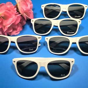 May include: Five white plastic sunglasses with black lenses. Each pair has a different name written on the right lens: Joni, Rhonnique, Donna, Woodell, and Roq.