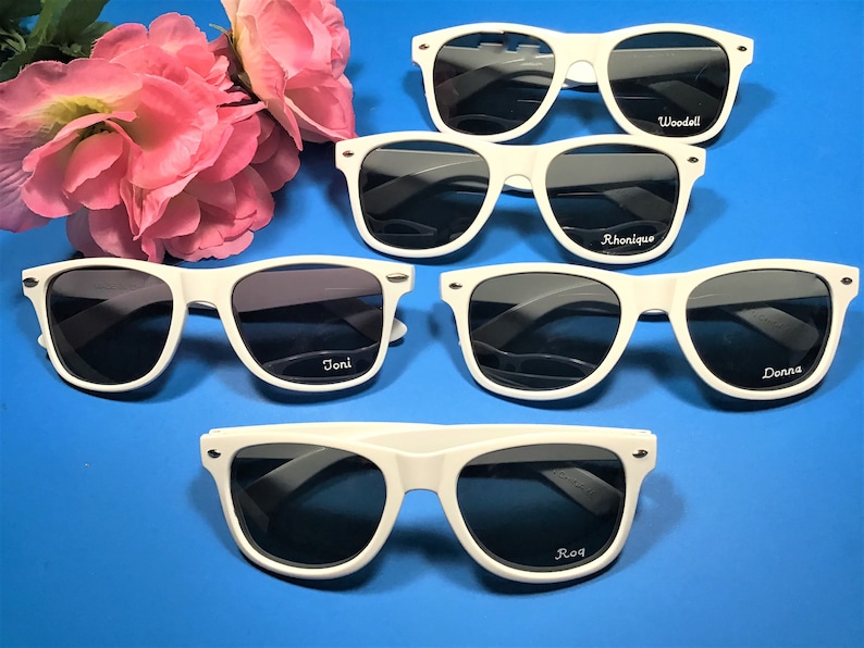May include: Five white plastic sunglasses with black lenses. Each pair has a different name written on the right lens: Joni, Rhonnique, Donna, Woodell, and Roq.