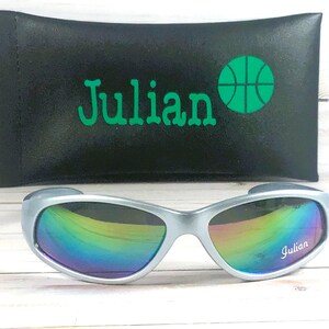 Op de afbeelding: Zilverkleurige zonnebril met iriserende, veelkleurige glazen met de naam Julian in het wit op het rechterglas. Achter de zonnebril ligt een zwart rechthoekig etui met de naam Julian en een groen basketbalpictogram.