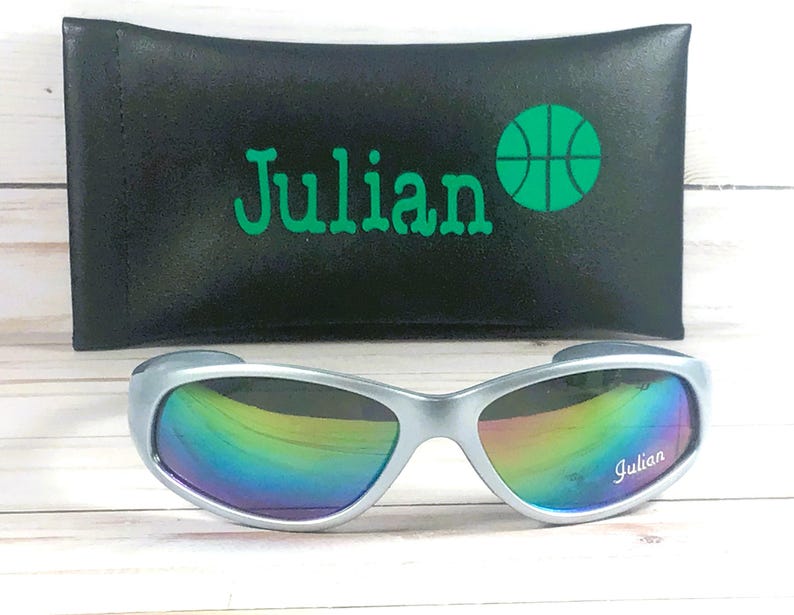 Op de afbeelding: Zilverkleurige zonnebril met iriserende, veelkleurige glazen met de naam Julian in het wit op het rechterglas. Achter de zonnebril ligt een zwart rechthoekig etui met de naam Julian en een groen basketbalpictogram.
