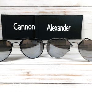 May include: Two pairs of aviator sunglasses with mirrored lenses. The sunglasses are on a white wooden surface. The sunglasses are in their cases, which are black with white text. The text on the cases reads "Cannon" and "Alexander".