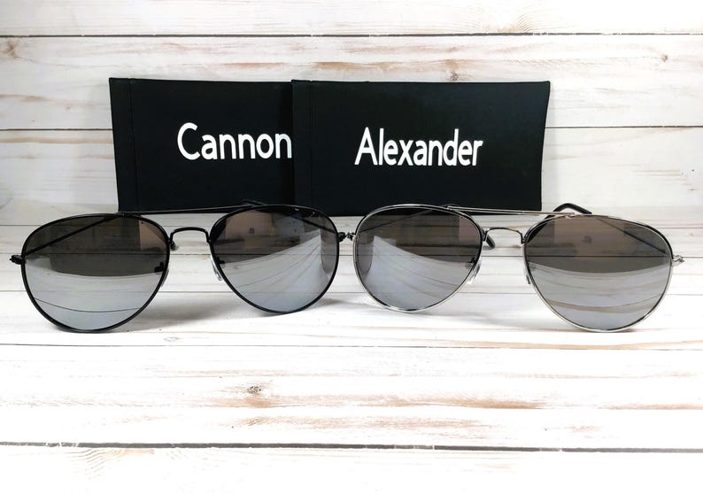 May include: Two pairs of aviator sunglasses with mirrored lenses. The sunglasses are on a white wooden surface. The sunglasses are in their cases, which are black with white text. The text on the cases reads "Cannon" and "Alexander".