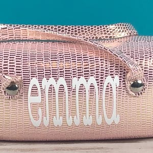 May include: A pink, textured handbag with a handle and the name "emma" in white script. The bag has silver-coloured hardware and a rectangular shape. The background is a light blue.