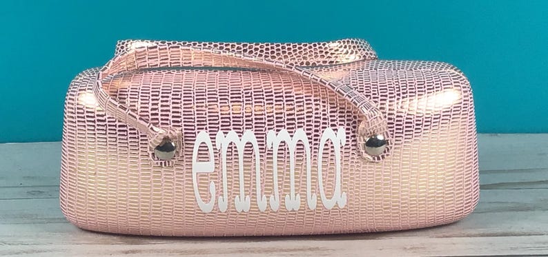 May include: A pink, textured handbag with a handle and the name "emma" in white script. The bag has silver-coloured hardware and a rectangular shape. The background is a light blue.