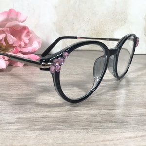 Puede incluir: Gafas con montura negra con lentes transparentes y pedrer&iacute;a rosa decorativa en las esquinas superiores. Las gafas tienen un puente y patillas plateadas. Una flor rosa est&aacute; en el fondo.