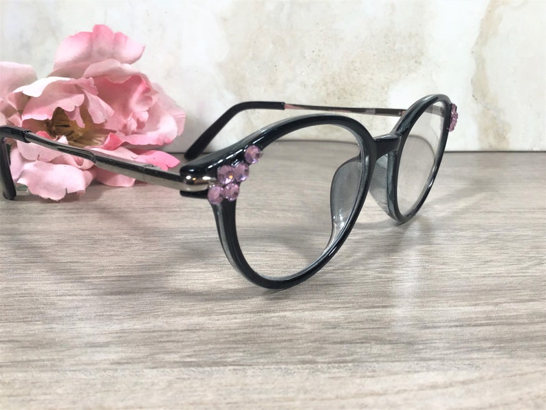 Puede incluir: Gafas con montura negra con lentes transparentes y pedrer&iacute;a rosa decorativa en las esquinas superiores. Las gafas tienen un puente y patillas plateadas. Una flor rosa est&aacute; en el fondo.