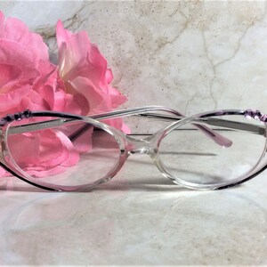 Puede incluir: Gafas transparentes con montura morada y plateada. Las gafas tienen detalles decorativos en las patillas y est&aacute;n colocadas frente a flores rosas. Las lentes son de forma ovalada.