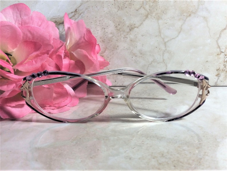 Puede incluir: Gafas transparentes con montura morada y plateada. Las gafas tienen detalles decorativos en las patillas y est&aacute;n colocadas frente a flores rosas. Las lentes son de forma ovalada.