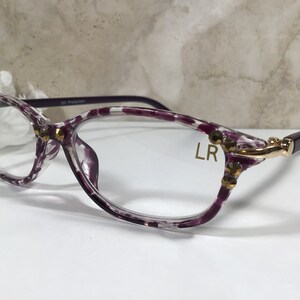 Puede incluir: Gafas transparentes con montura con estampado de lunares morados y blancos. Las monturas est&aacute;n adornadas con adornos dorados. Las patillas son de color morado oscuro. El texto "LR" est&aacute; impreso en la lente.