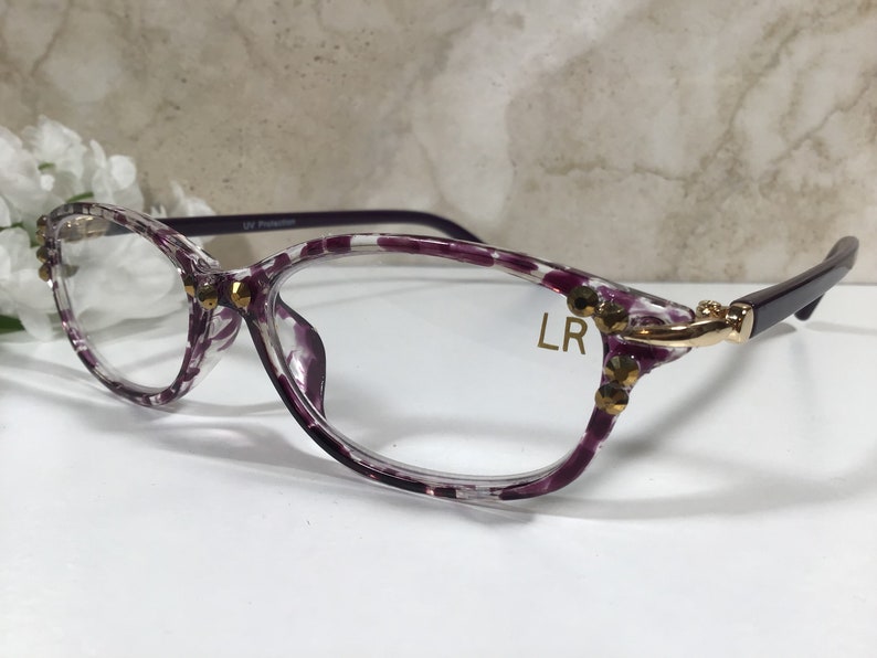 Puede incluir: Gafas transparentes con montura con estampado de lunares morados y blancos. Las monturas est&aacute;n adornadas con adornos dorados. Las patillas son de color morado oscuro. El texto "LR" est&aacute; impreso en la lente.